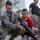 LCF-24 and RS Fort Lauderdale Poolee Function A U.S. Marine in digital camouflage uniform is demonstrating a firearm to a young civilian wearing a red shirt and glasses. The setting appears to be an outdoor military facility or base, with shipping containers and a corrugated metal building visible in the background. Additional Marines and civilians are visible in the background, suggesting a public outreach or recruitment event.