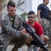 A U.S. Marine in digital camouflage uniform is demonstrating a firearm to a young civilian wearing a red shirt and glasses. The setting appears to be an outdoor military facility or base, with shipping containers and a corrugated metal building visible in the background. Additional Marines and civilians are visible in the background, suggesting a public outreach or recruitment event.