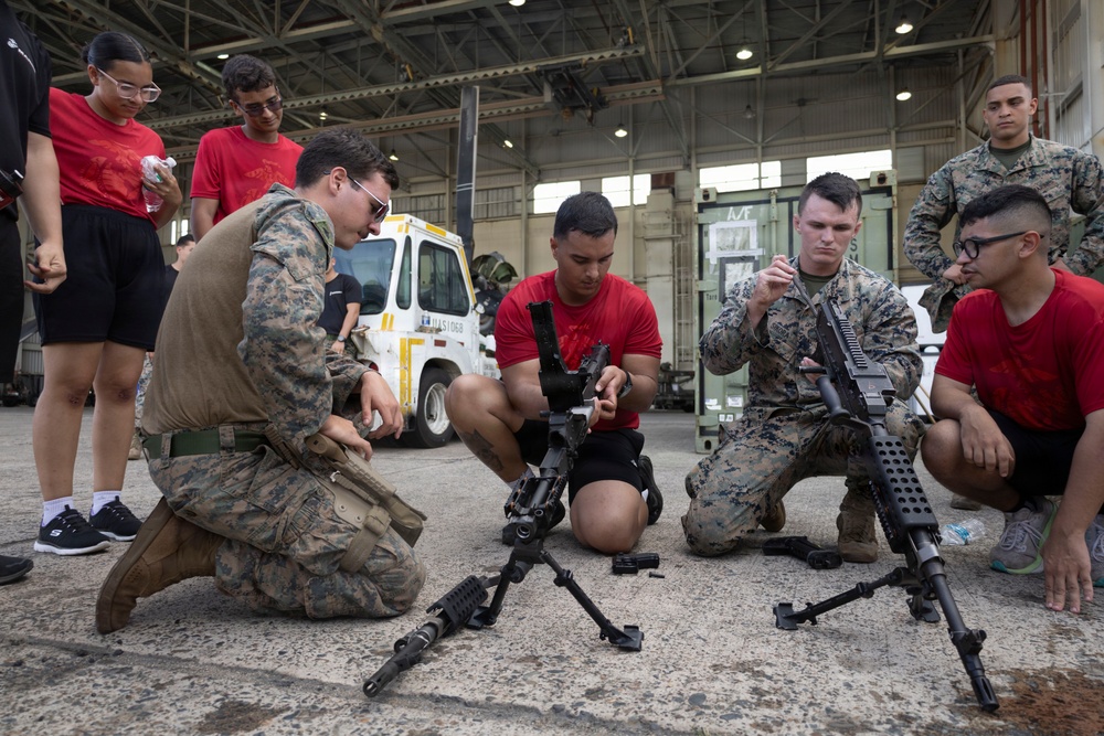 Two U.S. Marines in digital camouflage uniforms are conducting a weapons familiarization demonstration inside a military hangar facility. They are showing disassembled rifles on bipods to a group of young civilians wearing red shirts, likely participants in a youth outreach or JROTC-type program. The setting appears to be an official military open house or educational event.