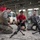 LCF-24 and RS Fort Lauderdale Poolee Function Two U.S. Marines in digital camouflage uniforms are conducting a weapons familiarization demonstration inside a military hangar facility. They are showing disassembled rifles on bipods to a group of young civilians wearing red shirts, likely participants in a youth outreach or JROTC-type program. The setting appears to be an official military open house or educational event.
