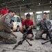 Two U.S. Marines in digital camouflage uniforms are conducting a weapons familiarization demonstration inside a military hangar facility. They are showing disassembled rifles on bipods to a group of young civilians wearing red shirts, likely participants in a youth outreach or JROTC-type program. The setting appears to be an official military open house or educational event.