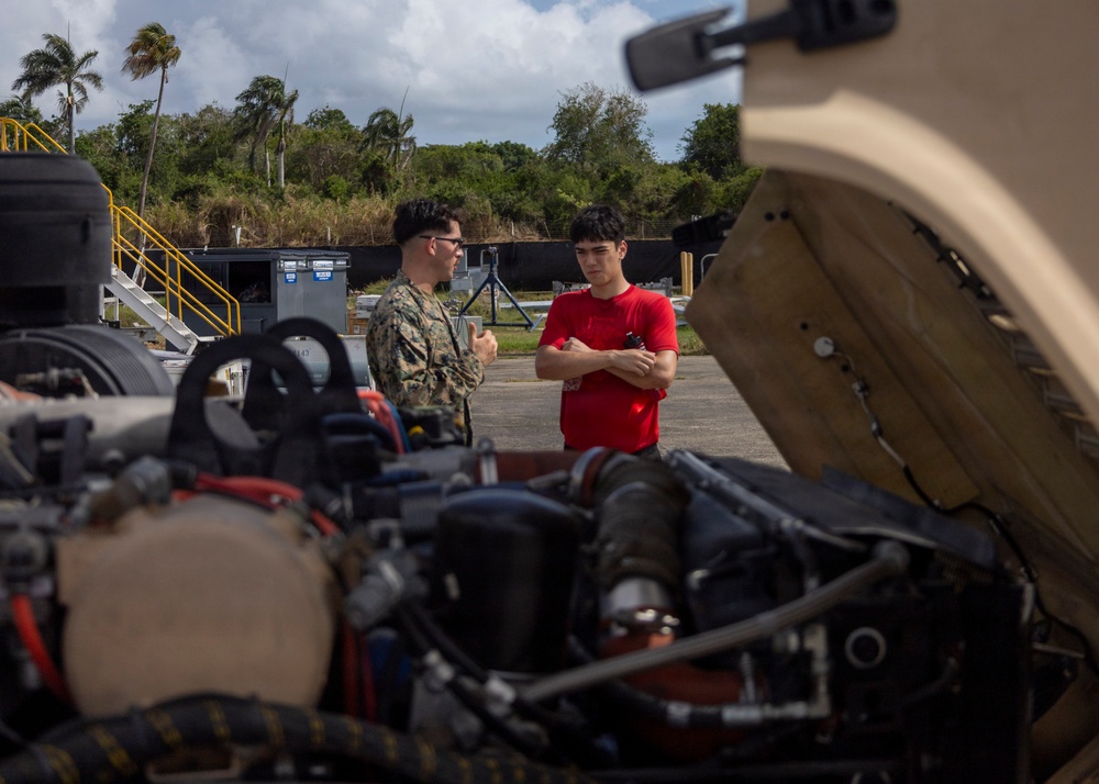Two individuals stand near an open-hood military vehicle, appearing to discuss mechanical work or maintenance. One person is wearing military camouflage uniform while the other is in a red civilian t-shirt with arms crossed. The setting appears to be an outdoor military installation or motor pool, with tropical vegetation and support equipment visible in the background.