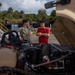 Two individuals stand near an open-hood military vehicle, appearing to discuss mechanical work or maintenance. One person is wearing military camouflage uniform while the other is in a red civilian t-shirt with arms crossed. The setting appears to be an outdoor military installation or motor pool, with tropical vegetation and support equipment visible in the background.