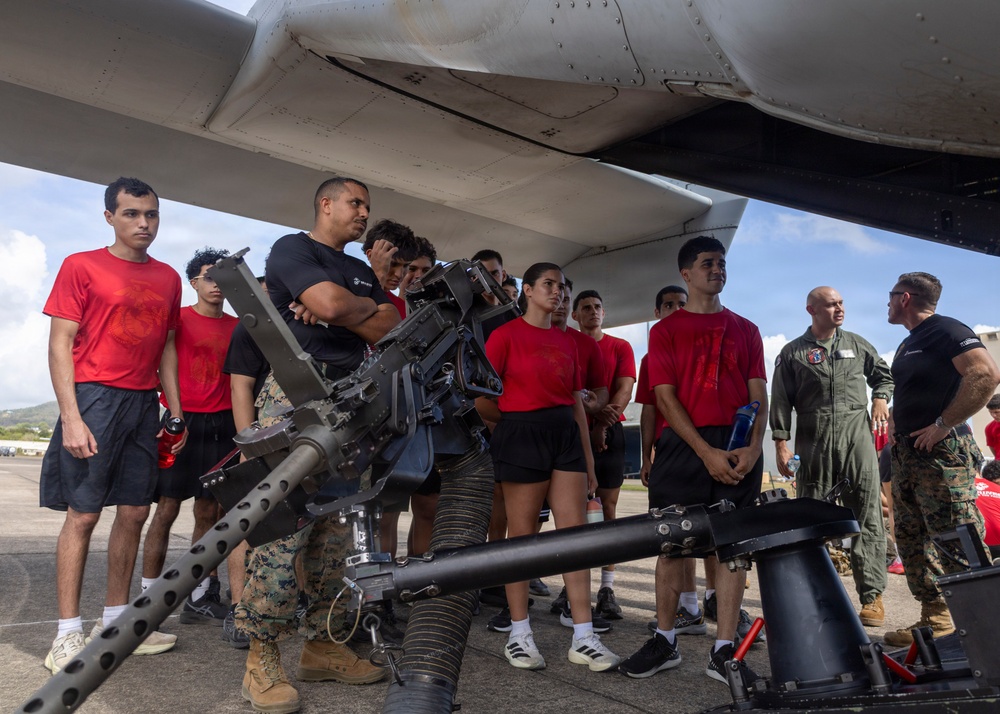 A group of young people wearing red t-shirts and athletic wear are gathered under the fuselage of a military aircraft, receiving a briefing or demonstration from uniformed military personnel. A large crew-served weapon system, appearing to be a .50 caliber machine gun or similar heavy weapon mounted on a stand, is prominently displayed in the foreground. The setting appears to be a military airfield or tarmac, suggesting this is a recruitment event, orientation, or educational demonstration.