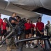 A group of young people wearing red t-shirts and athletic wear are gathered under the fuselage of a military aircraft, receiving a briefing or demonstration from uniformed military personnel. A large crew-served weapon system, appearing to be a .50 caliber machine gun or similar heavy weapon mounted on a stand, is prominently displayed in the foreground. The setting appears to be a military airfield or tarmac, suggesting this is a recruitment event, orientation, or educational demonstration.