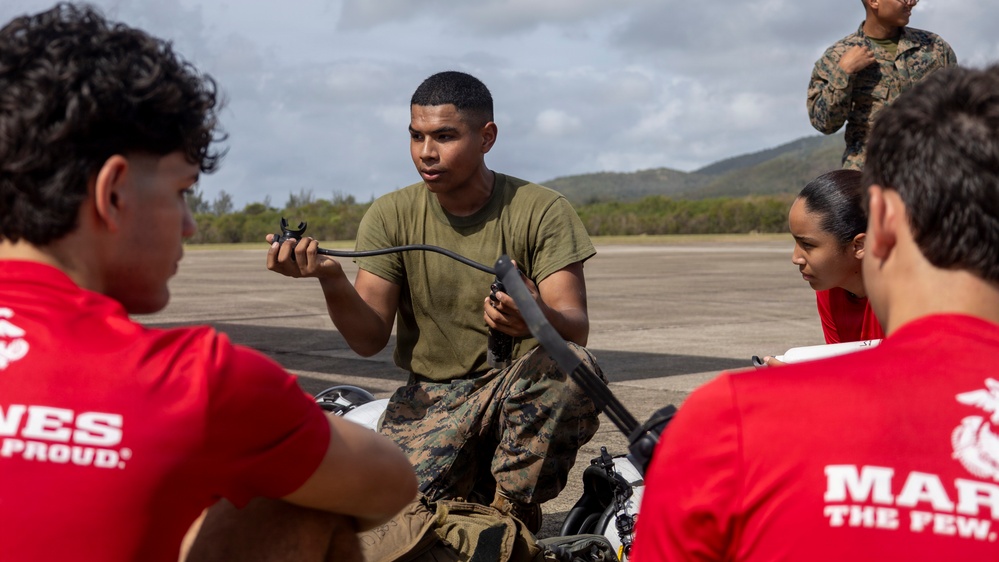 A U.S. Marine in combat utility uniform and olive drab t-shirt is conducting a demonstration or briefing for a group of young civilians wearing red 'Marines: The Few, The Proud' t-shirts. The Marine is holding what appears to be a component of a small unmanned aerial system (drone) and explaining it to the audience. The setting appears to be an outdoor tarmac or airstrip with hills visible in the background.
