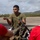 LCF-24 and RS Fort Lauderdale Poolee Function A U.S. Marine in combat utility uniform and olive drab t-shirt is conducting a demonstration or briefing for a group of young civilians wearing red 'Marines: The Few, The Proud' t-shirts. The Marine is holding what appears to be a component of a small unmanned aerial system (drone) and explaining it to the audience. The setting appears to be an outdoor tarmac or airstrip with hills visible in the background.