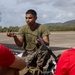 A U.S. Marine in combat utility uniform and olive drab t-shirt is conducting a demonstration or briefing for a group of young civilians wearing red 'Marines: The Few, The Proud' t-shirts. The Marine is holding what appears to be a component of a small unmanned aerial system (drone) and explaining it to the audience. The setting appears to be an outdoor tarmac or airstrip with hills visible in the background.