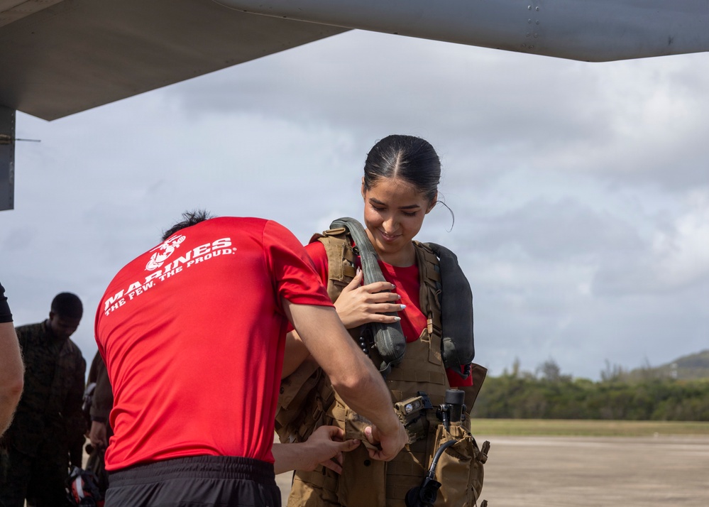 A Marine in a red 'Marines: The Few. The Proud.' PT shirt is assisting a young woman in fitting or adjusting flight/survival gear on an airfield tarmac. The setting appears to be a military airfield in a tropical location, with an aircraft visible overhead. The woman is smiling and appears to be a civilian or guest participant in a familiarization or orientation activity.