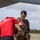 LCF-24 and RS Fort Lauderdale Poolee Function A Marine in a red 'Marines: The Few. The Proud.' PT shirt is assisting a young woman in fitting or adjusting flight/survival gear on an airfield tarmac. The setting appears to be a military airfield in a tropical location, with an aircraft visible overhead. The woman is smiling and appears to be a civilian or guest participant in a familiarization or orientation activity.