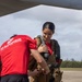 A Marine in a red 'Marines: The Few. The Proud.' PT shirt is assisting a young woman in fitting or adjusting flight/survival gear on an airfield tarmac. The setting appears to be a military airfield in a tropical location, with an aircraft visible overhead. The woman is smiling and appears to be a civilian or guest participant in a familiarization or orientation activity.