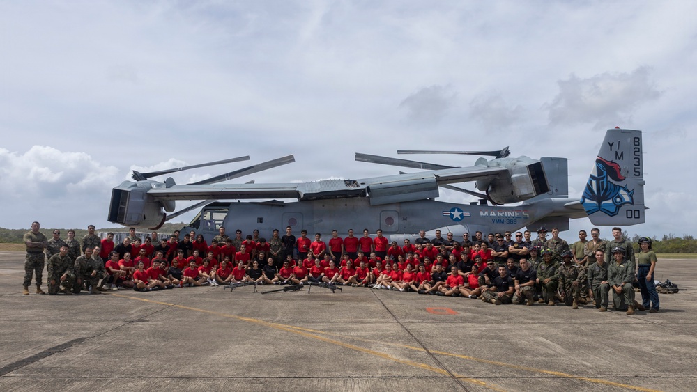A large group of military personnel and civilians in red shirts pose for a group photograph in front of a U.S. Marines MV-22 Osprey tiltrotor aircraft on a tarmac. The aircraft displays the tail code 'YM' and number '8235', along with a unit insignia on the tail fin, identifying it as belonging to VMM-265. The setting appears to be an outdoor airfield with overcast skies and tropical vegetation visible in the background.