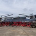 A large group of military personnel and civilians in red shirts pose for a group photograph in front of a U.S. Marines MV-22 Osprey tiltrotor aircraft on a tarmac. The aircraft displays the tail code 'YM' and number '8235', along with a unit insignia on the tail fin, identifying it as belonging to VMM-265. The setting appears to be an outdoor airfield with overcast skies and tropical vegetation visible in the background.