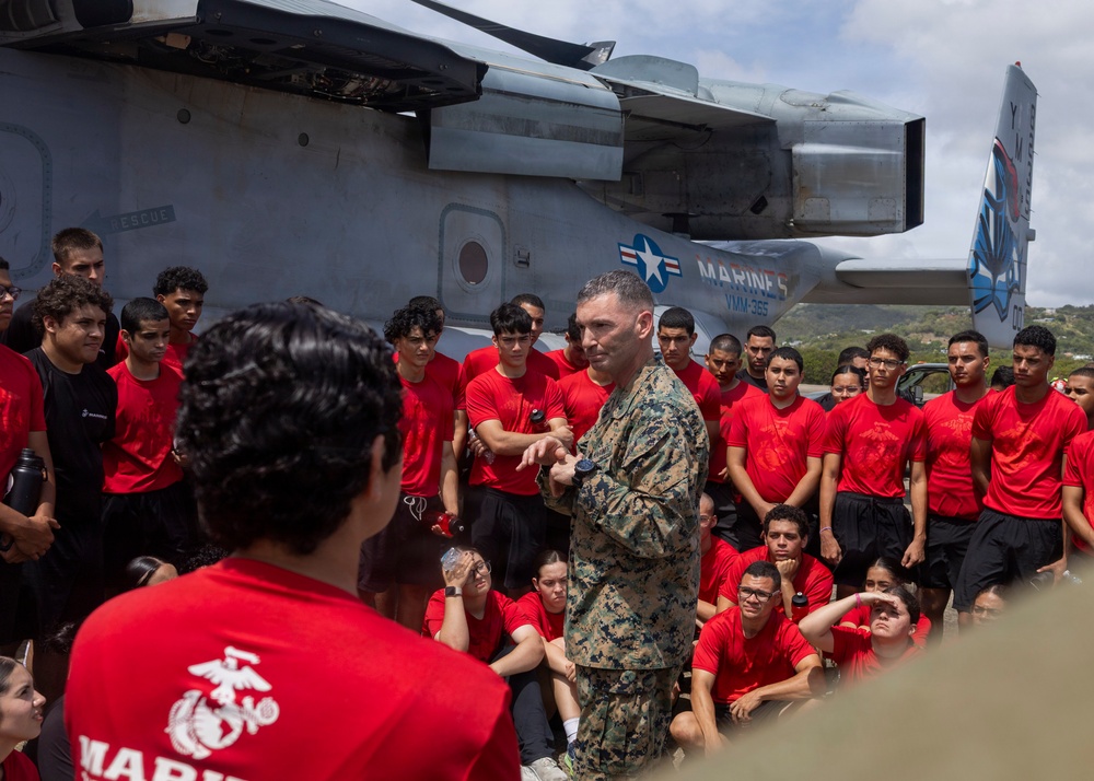 A U.S. Marine in camouflage uniform addresses a large group of young people wearing matching red 'Marines' t-shirts and black shorts, gathered in front of a military tiltrotor aircraft. The aircraft visible in the background is an MV-22 Osprey marked with 'VMM-365' squadron designation and U.S. Marines insignia. This appears to be a recruiting or youth outreach event conducted by the United States Marine Corps.