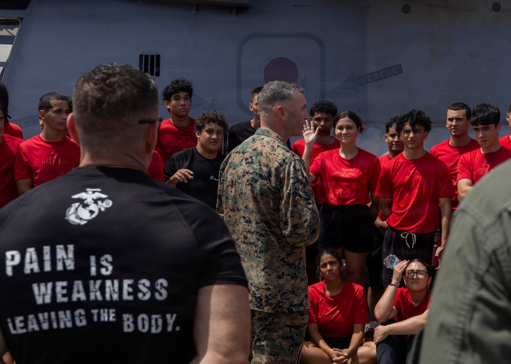 A U.S. Marine in camouflage uniform addresses a group of young people wearing red shirts, likely recruits or youth program participants, on what appears to be the deck of a naval vessel. A Marine in a black t-shirt reading 'Pain is Weakness Leaving the Body' stands in the foreground with his back to the camera. The setting suggests a military orientation, outreach, or recruiting event.