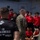 LCF-24 and RS Fort Lauderdale Poolee Function A U.S. Marine in camouflage uniform addresses a group of young people wearing red shirts, likely recruits or youth program participants, on what appears to be the deck of a naval vessel. A Marine in a black t-shirt reading 'Pain is Weakness Leaving the Body' stands in the foreground with his back to the camera. The setting suggests a military orientation, outreach, or recruiting event.