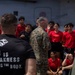 A U.S. Marine in camouflage uniform addresses a group of young people wearing red shirts, likely recruits or youth program participants, on what appears to be the deck of a naval vessel. A Marine in a black t-shirt reading 'Pain is Weakness Leaving the Body' stands in the foreground with his back to the camera. The setting suggests a military orientation, outreach, or recruiting event.