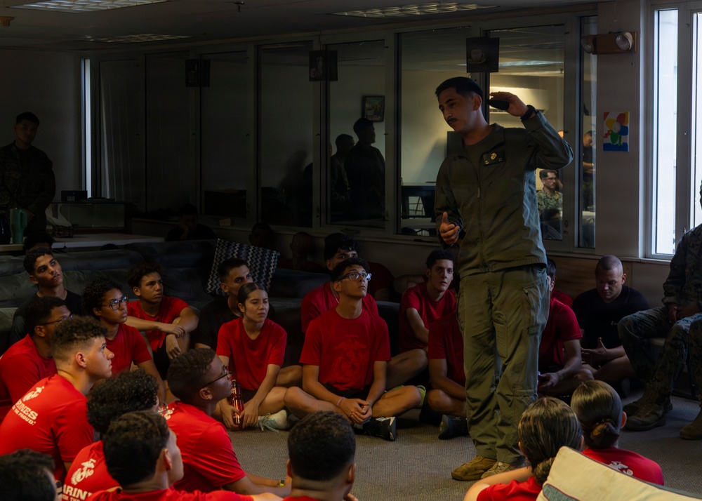 A military personnel member in olive/flight suit uniform addresses a group of young people seated on the floor, all wearing matching red t-shirts with what appears to be Marines branding. The setting appears to be an indoor briefing or orientation session, likely a recruiting or youth outreach event. Additional military personnel are visible in the background observing the presentation.