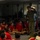 LCF-24 and RS Fort Lauderdale Poolee Function A military personnel member in olive/flight suit uniform addresses a group of young people seated on the floor, all wearing matching red t-shirts with what appears to be Marines branding. The setting appears to be an indoor briefing or orientation session, likely a recruiting or youth outreach event. Additional military personnel are visible in the background observing the presentation.