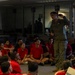 A military personnel member in olive/flight suit uniform addresses a group of young people seated on the floor, all wearing matching red t-shirts with what appears to be Marines branding. The setting appears to be an indoor briefing or orientation session, likely a recruiting or youth outreach event. Additional military personnel are visible in the background observing the presentation.