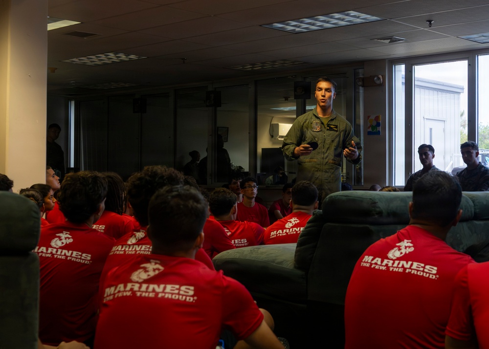 A military aviator in an olive green flight suit addresses a group of young people wearing red 'Marines - The Few, The Proud' t-shirts in an indoor setting. The speaker appears to be giving a presentation or briefing, likely as part of a Marine Corps recruitment or outreach event. The audience is seated and attentively facing the speaker.