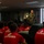 LCF-24 and RS Fort Lauderdale Poolee Function A military aviator in an olive green flight suit addresses a group of young people wearing red 'Marines - The Few, The Proud' t-shirts in an indoor setting. The speaker appears to be giving a presentation or briefing, likely as part of a Marine Corps recruitment or outreach event. The audience is seated and attentively facing the speaker.