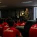 A military aviator in an olive green flight suit addresses a group of young people wearing red 'Marines - The Few, The Proud' t-shirts in an indoor setting. The speaker appears to be giving a presentation or briefing, likely as part of a Marine Corps recruitment or outreach event. The audience is seated and attentively facing the speaker.