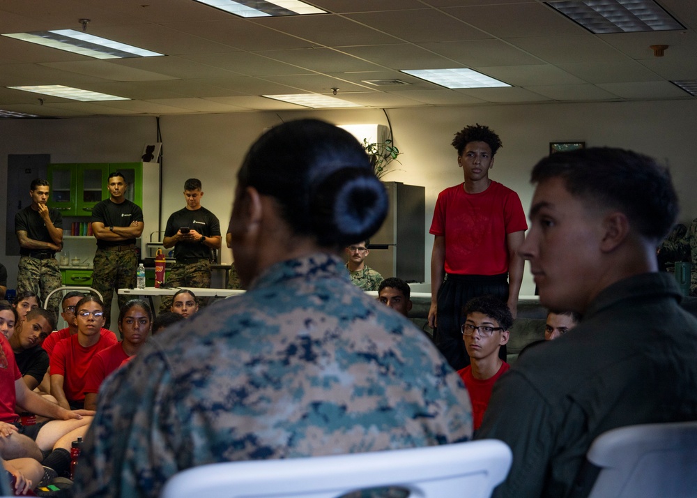 A military briefing or orientation session is taking place indoors, with uniformed Marines addressing a group of young civilians wearing red shirts. The scene appears to be a youth outreach or recruitment event, possibly a Junior ROTC or similar program. A Marine in camouflage uniform is speaking to seated participants while others stand and observe.