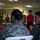 LCF-24 and RS Fort Lauderdale Poolee Function A military briefing or orientation session is taking place indoors, with uniformed Marines addressing a group of young civilians wearing red shirts. The scene appears to be a youth outreach or recruitment event, possibly a Junior ROTC or similar program. A Marine in camouflage uniform is speaking to seated participants while others stand and observe.
