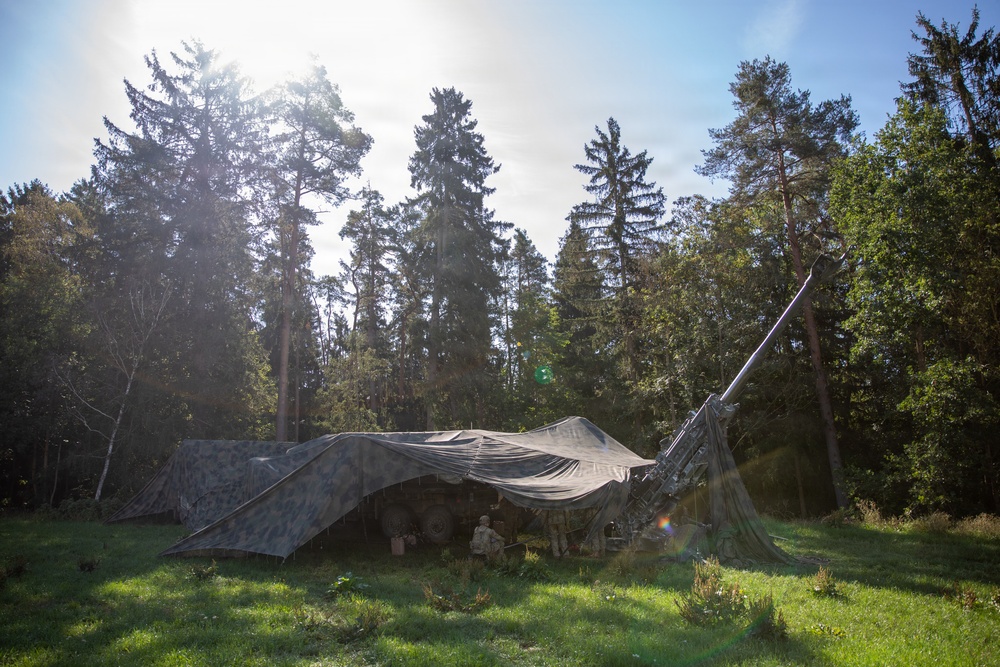 A military field camp is set up in a forest clearing, covered by a large camouflage netting to conceal it from aerial observation. A tall antenna or mast structure is visible extending upward on the right side of the camp. At least one soldier appears to be present beneath the netting near military equipment or vehicles.