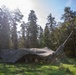 A military field camp is set up in a forest clearing, covered by a large camouflage netting to conceal it from aerial observation. A tall antenna or mast structure is visible extending upward on the right side of the camp. At least one soldier appears to be present beneath the netting near military equipment or vehicles.
