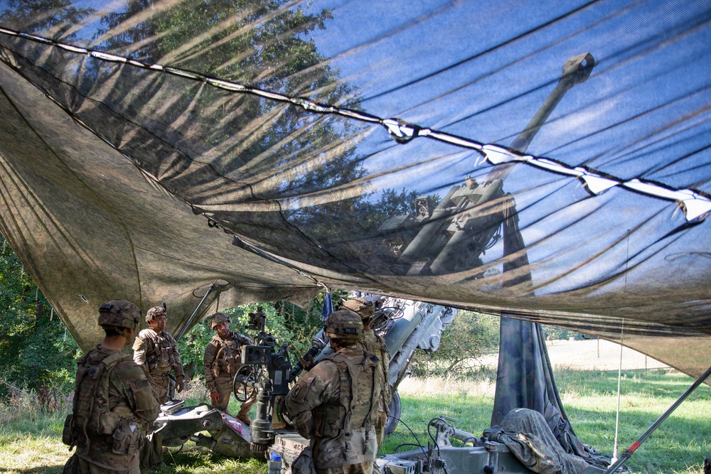 Military personnel in camouflage uniforms are gathered around what appears to be an artillery piece, likely an M777 howitzer or similar towed artillery system, positioned under a large camouflage netting. The scene takes place in a field or wooded area, with the netting providing concealment from aerial observation. A second artillery piece is visible in the background through the semi-transparent netting.