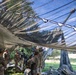 Military personnel in camouflage uniforms are gathered around what appears to be an artillery piece, likely an M777 howitzer or similar towed artillery system, positioned under a large camouflage netting. The scene takes place in a field or wooded area, with the netting providing concealment from aerial observation. A second artillery piece is visible in the background through the semi-transparent netting.