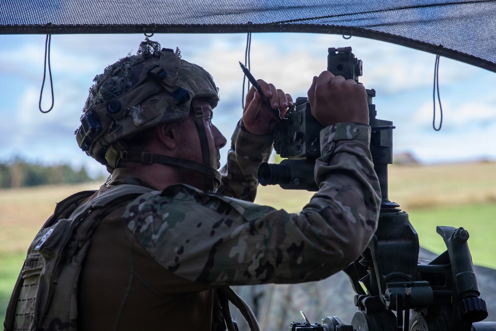 A soldier in full combat gear is operating what appears to be an artillery or mortar sighting/aiming device under a camouflage net. The soldier is wearing MultiCam (OCP) pattern uniform, a tactical helmet with accessories, and a plate carrier vest. The setting appears to be an outdoor field environment, likely during a military training exercise.