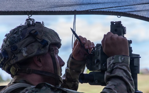 A soldier in full combat gear is operating what appears to be an artillery or mortar sighting/aiming device under a camouflage net. The soldier is wearing MultiCam (OCP) pattern uniform, a tactical helmet with accessories, and a plate carrier vest. The setting appears to be an outdoor field environment, likely during a military training exercise.