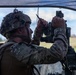 A soldier in full combat gear is operating what appears to be an artillery or mortar sighting/aiming device under a camouflage net. The soldier is wearing MultiCam (OCP) pattern uniform, a tactical helmet with accessories, and a plate carrier vest. The setting appears to be an outdoor field environment, likely during a military training exercise.