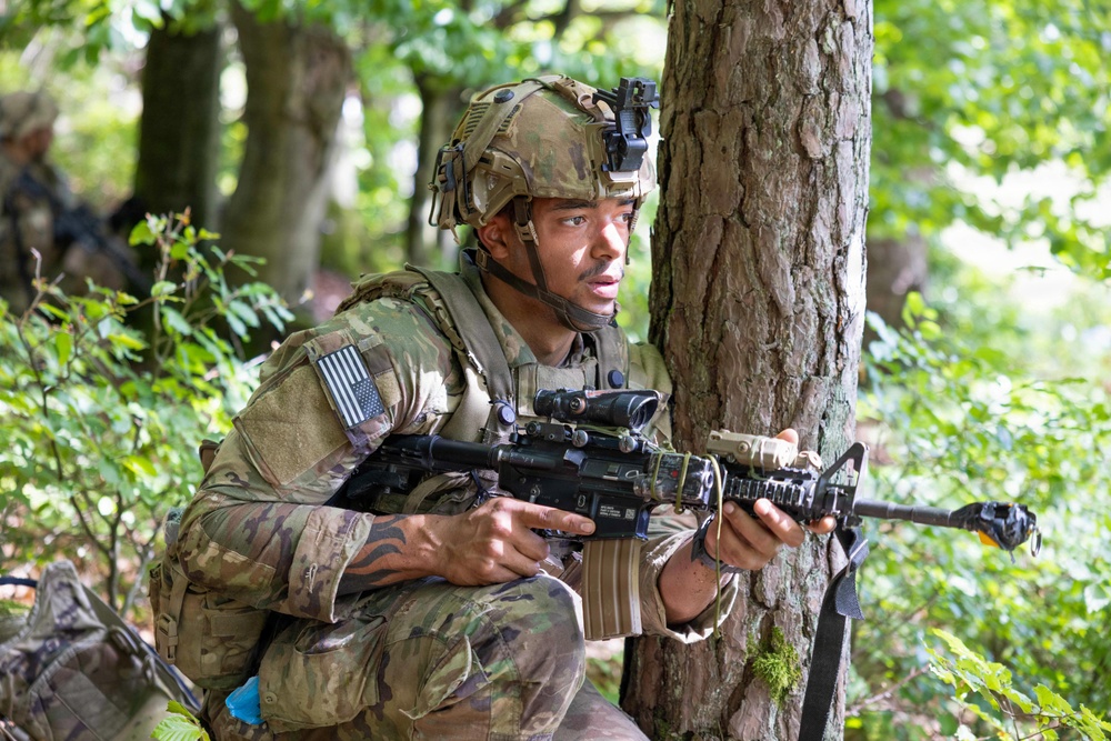 A U.S. Army soldier in full combat gear is positioned behind a tree in a wooded area, appearing to be in a tactical training exercise or field operation. The soldier is wearing MultiCam camouflage, a tactical helmet with night vision mount, and is holding an M4-style carbine rifle. An American flag patch is visible on the left shoulder of the uniform.