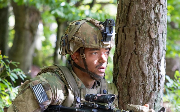A U.S. Army soldier in full combat gear is positioned behind a tree in a wooded area, appearing to be in a tactical training exercise or field operation. The soldier is wearing MultiCam camouflage, a tactical helmet with night vision mount, and is holding an M4-style carbine rifle. An American flag patch is visible on the left shoulder of the uniform.