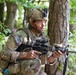 A U.S. Army soldier in full combat gear is positioned behind a tree in a wooded area, appearing to be in a tactical training exercise or field operation. The soldier is wearing MultiCam camouflage, a tactical helmet with night vision mount, and is holding an M4-style carbine rifle. An American flag patch is visible on the left shoulder of the uniform.
