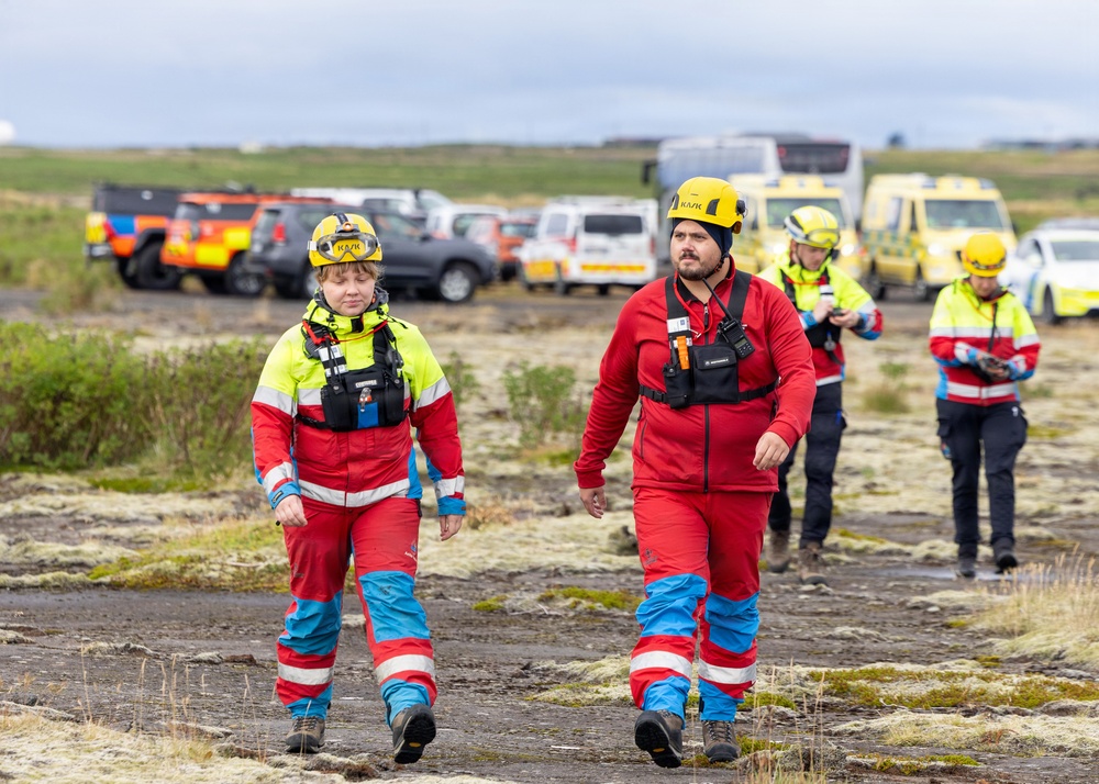 A group of search and rescue personnel are walking across a rugged, dark terrain that appears to be volcanic landscape. They are wearing standardized red and blue uniforms with high-visibility yellow jackets, yellow helmets marked 'KASK', and radio communication equipment. Multiple emergency vehicles including ambulances are staged in the background, suggesting an active rescue operation.