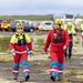 A group of search and rescue personnel are walking across a rugged, dark terrain that appears to be volcanic landscape. They are wearing standardized red and blue uniforms with high-visibility yellow jackets, yellow helmets marked 'KASK', and radio communication equipment. Multiple emergency vehicles including ambulances are staged in the background, suggesting an active rescue operation.
