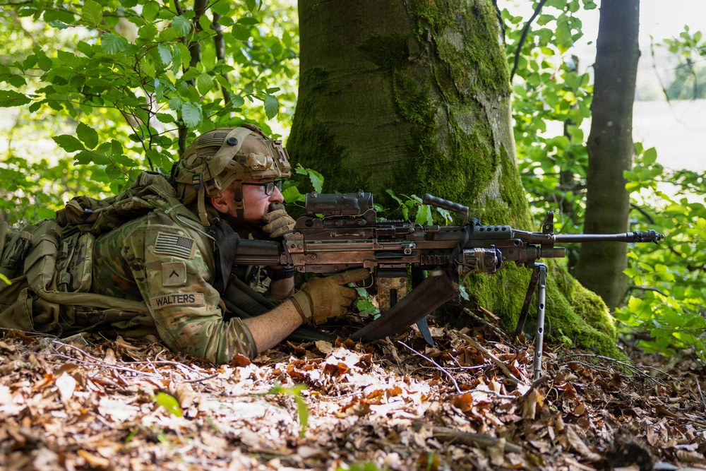 A U.S. Army soldier identified as 'Walters' is in a prone firing position in a wooded area, aiming a light machine gun supported by a bipod. The soldier is wearing full multicam combat gear including a tactical helmet, body armor, and a large rucksack. The setting appears to be a training exercise in a European forest environment.