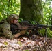 A U.S. Army soldier identified as 'Walters' is in a prone firing position in a wooded area, aiming a light machine gun supported by a bipod. The soldier is wearing full multicam combat gear including a tactical helmet, body armor, and a large rucksack. The setting appears to be a training exercise in a European forest environment.