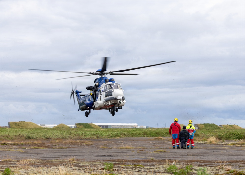 A large blue and white helicopter, appearing to be an Airbus H225 (Super Puma) type, is hovering low over a flat tarmac or airfield surface. Three individuals in high-visibility and emergency response gear are standing nearby observing the aircraft. The setting appears to be a coastal or remote airfield with grass mounds and industrial structures visible in the background.