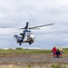 A large blue and white helicopter, appearing to be an Airbus H225 (Super Puma) type, is hovering low over a flat tarmac or airfield surface. Three individuals in high-visibility and emergency response gear are standing nearby observing the aircraft. The setting appears to be a coastal or remote airfield with grass mounds and industrial structures visible in the background.