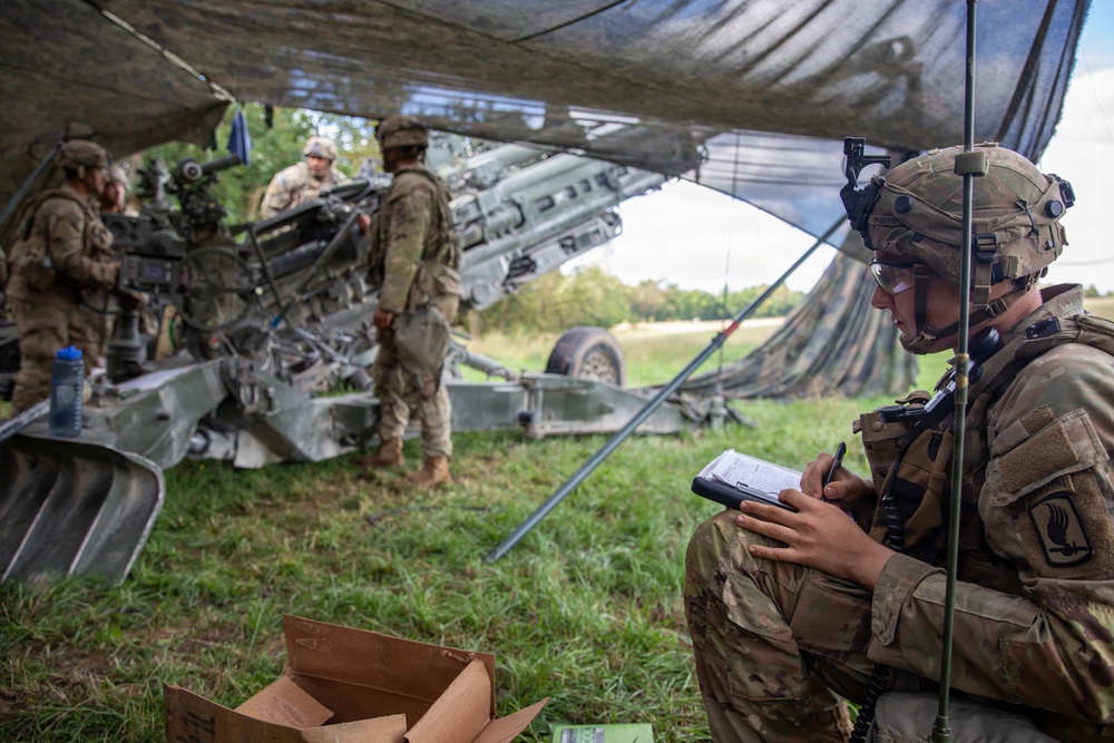 Military personnel are operating under a camouflage netting in a field setting, likely during a training exercise or field operation. A soldier in the foreground is seated and writing on a clipboard while wearing a helmet with communications equipment. In the background, several soldiers are working around what appears to be an M777 howitzer artillery piece.