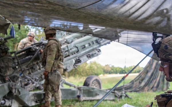 Military personnel are operating under a camouflage netting in a field setting, likely during a training exercise or field operation. A soldier in the foreground is seated and writing on a clipboard while wearing a helmet with communications equipment. In the background, several soldiers are working around what appears to be an M777 howitzer artillery piece.