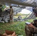 Military personnel are operating under a camouflage netting in a field setting, likely during a training exercise or field operation. A soldier in the foreground is seated and writing on a clipboard while wearing a helmet with communications equipment. In the background, several soldiers are working around what appears to be an M777 howitzer artillery piece.