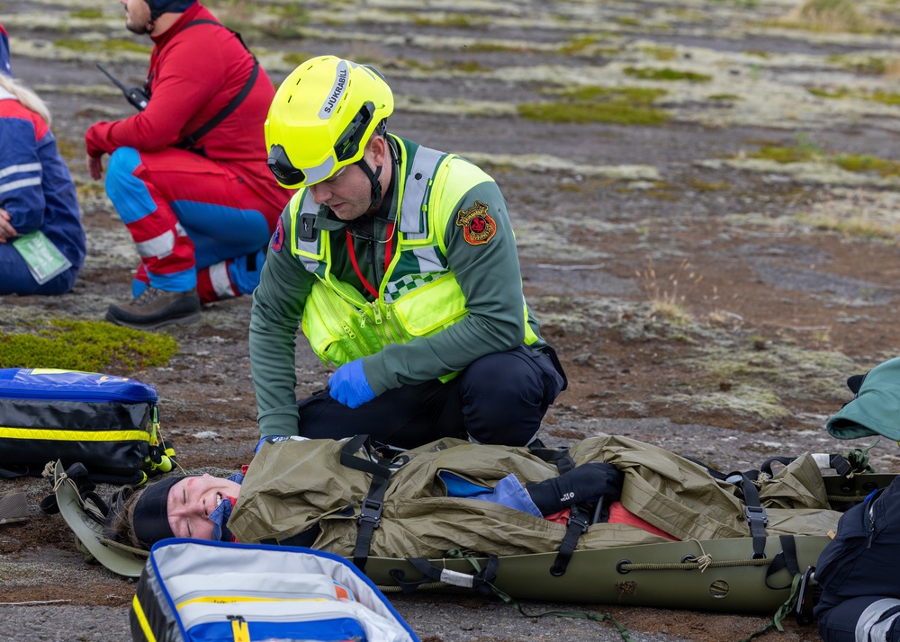 A rescue worker wearing a yellow helmet and high-visibility vest is attending to a patient strapped to an olive-green stretcher/backboard on the ground. The responder is wearing blue medical gloves and appears to be assessing or treating the patient. Additional emergency personnel in red and blue uniforms are visible in the background, suggesting a multi-casualty rescue exercise or incident.