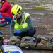 A rescue worker wearing a yellow helmet and high-visibility vest is attending to a patient strapped to an olive-green stretcher/backboard on the ground. The responder is wearing blue medical gloves and appears to be assessing or treating the patient. Additional emergency personnel in red and blue uniforms are visible in the background, suggesting a multi-casualty rescue exercise or incident.